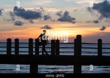 Ein Fotograf mit Kamera auf einem Stativ in der Silhouette bei Sonnenuntergang steht auf einer Plattform auf die Meerenge Melaka Masjid Al Hus neben korrigiert Stockfoto