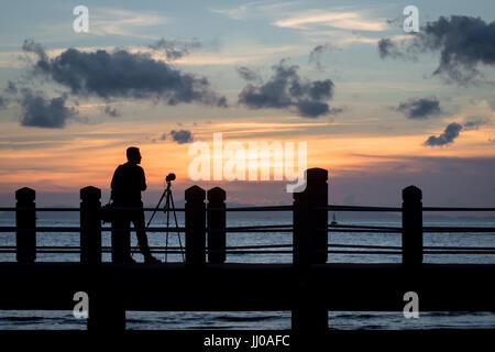 Ein Fotograf mit Kamera auf einem Stativ in der Silhouette bei Sonnenuntergang steht auf einer Plattform auf die Meerenge Melaka Masjid Al Hus neben korrigiert Stockfoto