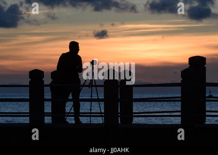 Ein Fotograf mit Kamera auf einem Stativ in der Silhouette bei Sonnenuntergang steht auf einer Plattform auf die Meerenge Melaka Masjid Al Hus neben korrigiert Stockfoto