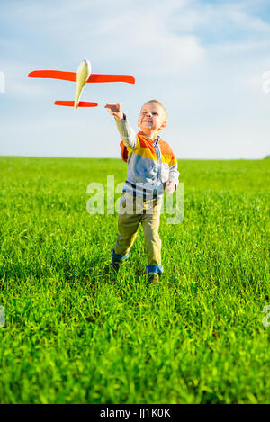 Fröhlicher Junge spielt mit Spielzeugflugzeug gegen blauen Himmel und grüne Feld Hintergrund. Stockfoto