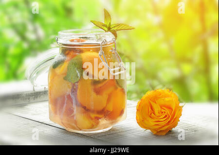 Kanne mit Deckel mit Wasser, detox mit Stücken von Pfirsich und Aprikose, mit Eis. Ein erfrischendes Getränk auf einem grünen Hintergrund mit einer gelben Rose. Stockfoto