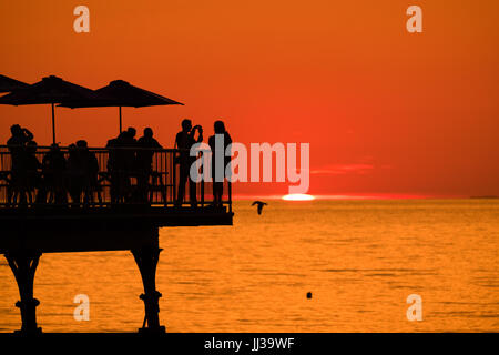 Aberystwyth Wales UK, Montag, 17. Juli 2017 UK Wetter: Menschen auf Aberystwyth Pier sind in den lebendigen goldenen Sonnenuntergang über den ruhigen Gewässern der Cardigan Bay, Silhouette, wie die Mini-Hitzewelle beginnt, über das UK Foto Kredit bauen: Keith Morris/Alamy Live News Stockfoto