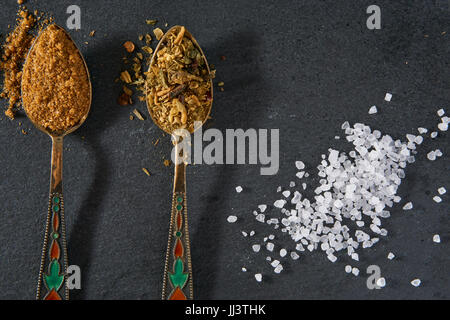 zwei kleine dekorative Löffel Gewürz Gewürze und Prise Meersalz auf schwarzem Schiefer. obere Winkel Detailansicht Stockfoto