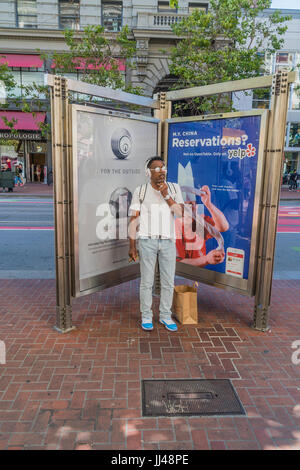 Eine afrikanisch-amerikanischen männlichen Pendler steht und wartet vor einer zweiseitigen Plakatwand an der Market Street in San Francisco. Er trägt reflektierenden Spiegel-lik Stockfoto