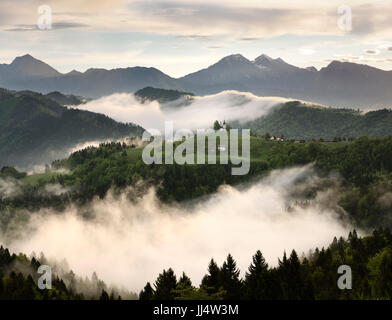 Sanften Nebel bei Sonnenaufgang mit Bergen von Kamnik Savinja Alpen Skofjelosko Hills mit St. Thomaskirche in der Nähe von Ljubljana Slowenien Stockfoto