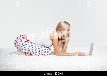 Junge blonde Frau im Schlafanzug auf weißen Laptop Surfen gesamte-Teppich sitzen Stockfoto