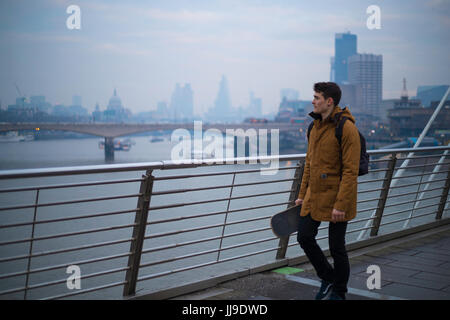 Ein junger Mann kreuzt die Jubilee Bridge in London mit dem Skateboard in seiner Hand. Stockfoto