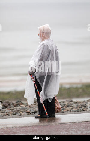 Eine ältere Frau allein stehend auf der Küstenpromenade in Llandudno in einem durchsichtigen Kunststoff Poncho mit Kapuze, Blick auf das Meer, wie sie tief in Gedanken, Llandudno, Wales, Vereinigtes Königreich von den sintflutartigen Regen Regen nass wird Stockfoto