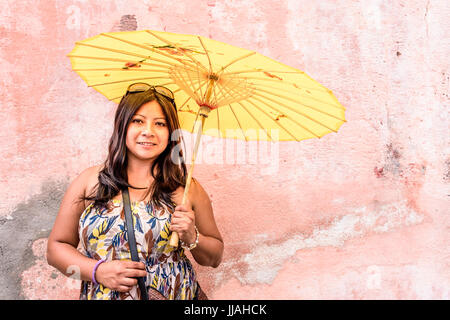Porträt der jungen Hispanic Frau im Sommerkleid holding Gelb im chinesischen Stil Sonnenschirm & stehend gegen alte, abgenutzte, verblasste, rosa Wand Stockfoto