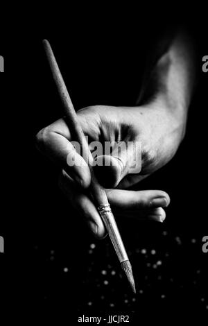 Die Hände des Künstlers halten Sie eine Bürste. auf einem schwarzen Hintergrund mit tiefen Schatten und hellen Highlights. Schwarze und Weiße konzeptionelle Bild Stockfoto