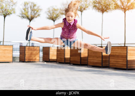 Aktives Kind Mädchen Turnerin springen auf der Straße tun spaltet. Stretching-Übung, gerade Winkel Pose in der Luft. Junges Mädchen Acrobat. Das Mädchen beschäftigt sich mit Gymnastik. Stockfoto