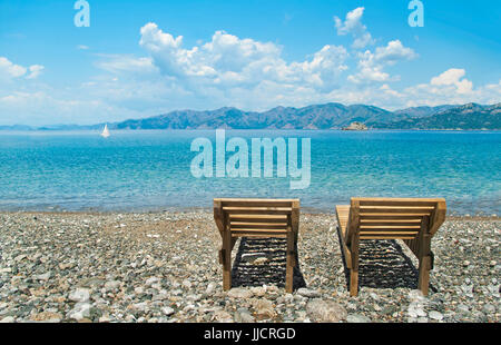 zwei hölzerne Sonnenliegen am Kiesstrand mit Blick auf Berge, blauer Himmel, Wolken und weiße Segelboot, Marmaris, Türkei Stockfoto