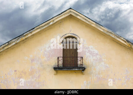 mit Brettern vernagelt Balkon auf verwitterten verputzte Wand des Giebels mit abblätternde Farbe. altes verlassenes Haus gegen dunkel bewölkter Himmel. Stockfoto