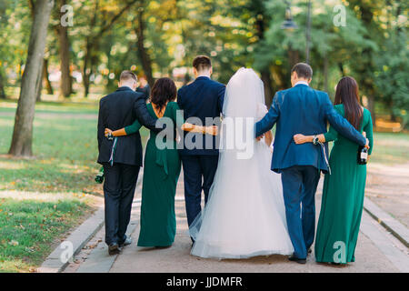 Die Rückansicht der das Brautpaar und ihre Frined im Park spazieren. Stockfoto