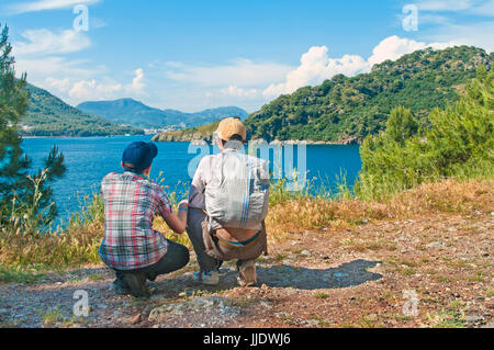 Kaukasische Teenager-Sohn in Karohemd und Mütze mit seinem Vater betrachten Marmaris Bucht Meer mit Bergen im Hintergrund am sonnigen Tag, Türkei Stockfoto