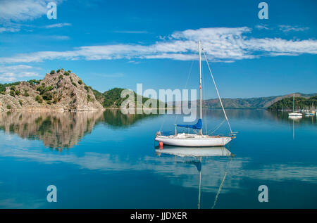weiße Yacht im Ägäischen Meer in der Nähe von felsigen Insel mit Bergen und Marti Marina am Hintergrund blauer Himmel mit weißen Wolken an sonnigen Tag in Orhaniye, Stockfoto