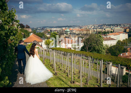 Die Rückansicht der stilvoll gekleidete Brautpaar händchenhaltend bei einem Spaziergang entlang der Pflasterung-Straße in der Nähe von Prag Panorama. Stockfoto