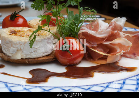 Gebratene Ziegenkäse mit Parmaschinken und Balsamico-Dressing auf einem weißen und blauen Teller Stockfoto