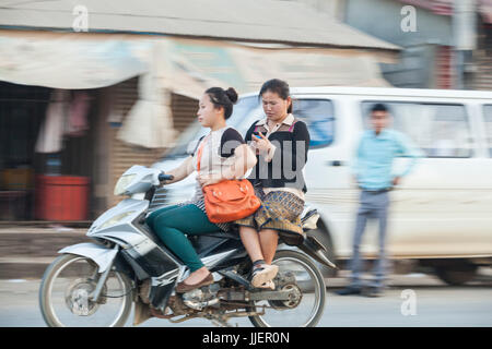 Junge Frauen fahren einen Roller zusammen und überprüfen Sie ihre Handys auf einer belebten Straße in Luang Prabang, Laos. Stockfoto