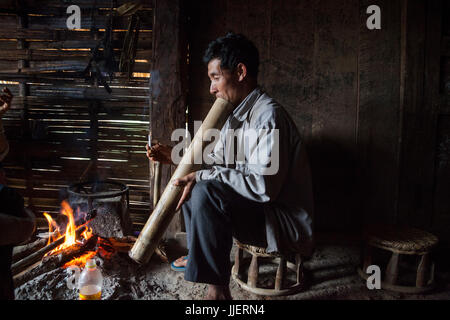 Ein Mann raucht eine Zigarette durch einen großen Bambus Wasser Bong durch einen Brand in einem Haus in Ban Sop-Kha, Laos. Stockfoto