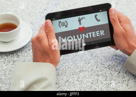 Unternehmer mit kleinen Tablet am Tisch gegen freiwillige Text mit verschiedenen Symbolen auf dem Bildschirm Stockfoto