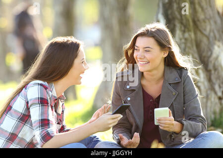 Zwei glückliche Freunde reden und lachen, halten ihre Smartphones sitzen auf dem Rasen in einem park Stockfoto