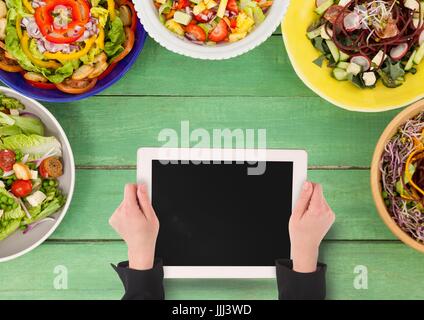 Person, die eine Tablette gegen einen grünen Holztisch mit Essen und Kopie Speicherplatz auf dem Handy Stockfoto