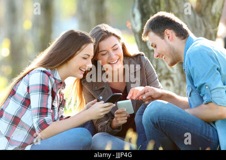 Drei Jugendliche Teilen auf Linie Inhalt auf ihren Smartphones sitzen auf dem Rasen in einem park Stockfoto