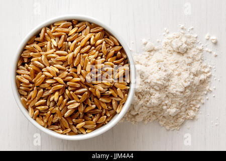 Kamut Weizenkörner in Keramikschale isoliert auf weiß lackiertem weißen Holz von oben. Neben weißem Mehl. Stockfoto