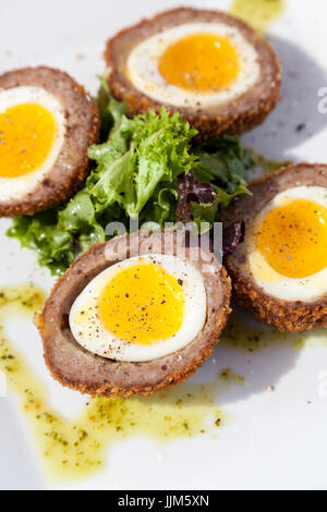 4 Dotter weichen Eiern umgeben von panierte und gebratene Wurst auf einem weißen Teller mit Beilagen und Nieselregen vorgestellt. Stockfoto