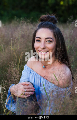 Eine wunderschöne junge Frau sitzt auf einer Wiese lange Gras in der Nähe von Liverpool eine Sommer Abend leichtes ziehen ihre Knie in Richtung Brust. Stockfoto