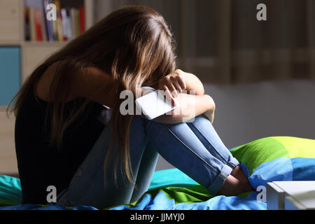 Einzige traurige Teen hält ein Handy beklagt sitzt auf dem Bett in ihrem Schlafzimmer mit einem dunklen Licht im Hintergrund Stockfoto