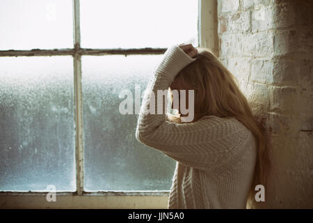 Seitenansicht einer miserablen jungen blonden Frau stehend durch die Fenster Kopf in Händen Stockfoto