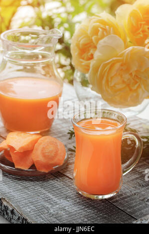 Karottensaft in Krug und Glas Klarglas auf einem Hintergrund von Rosen. Stockfoto