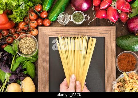 Draufsicht der Zusammensetzung von Lebensmitteln zu kochen Pasta zum Abendessen bereit. Menschliche Hände halten Nudeln über Blackboard mit Gemüse und Gewürzen um. Stockfoto