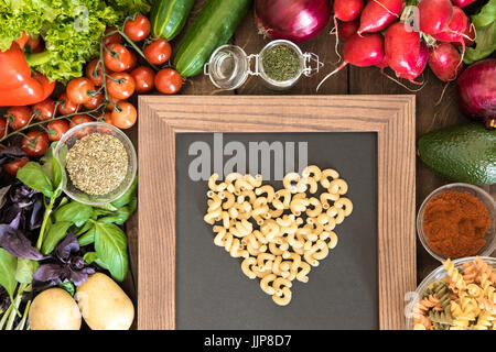 Kochen mit Liebe-Konzept. Küchentisch voller Gemüse, Gewürze und grün mit Herzschild von Pasta an Tafel gemacht, Stockfoto