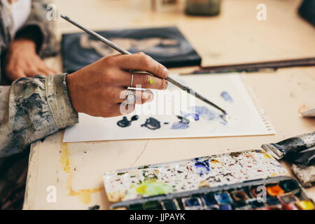 Nahaufnahme eines weiblichen Künstlers Hand hält Pinsel und Mischen von Farben auf Papier. Schuss der Malerin in ihrem Atelier arbeiten zugeschnitten. Stockfoto