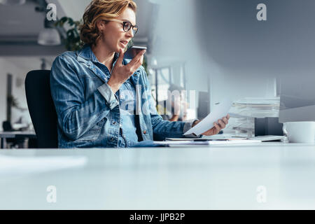 Junge Geschäftsfrau Lautsprecher telefonieren mit Dokument. Junge Frau sitzt im Büro mit Smartphone und mit Blick auf ein Papier. Stockfoto