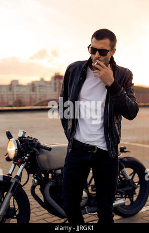 Schöne glücklicher Fahrer Mann mit Bart und Schnurrbart in schwarze Biker-Jacke, Jeans und Mode Sonnenbrillen Rauchen Luftbewegung in der Nähe von klassischen Stil Café Racer mo Stockfoto