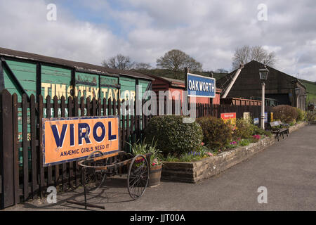 Blick auf alte Waggons, Anzeigen, Warenkorb, gas Lampen & verlassenen Plattform auf malerische, historische Oakworth Station - Keighley & Worth Valley Railway, England, UK. Stockfoto