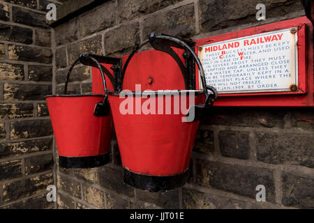 Anzeigen von 2 Old Red & black fire Eimer auf Plattform Wand an der malerischen, historischen Oakworth Station - Keighley & Worth Valley Railway, England, UK hängen. Stockfoto