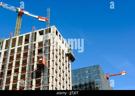 Niedrigen Winkel Blick auf ein konkretes Gebäude im Bau neben einem Glasbau mit zwei Turmdrehkrane gegen blauen Himmel. Stockfoto