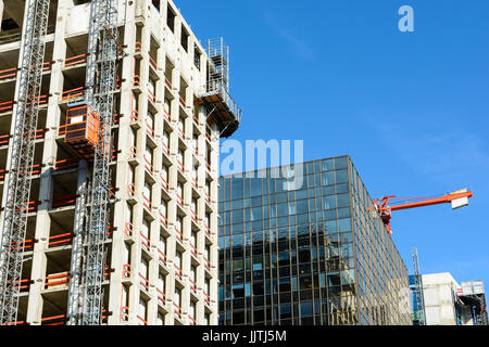 Niedrigen Winkel Blick auf ein Glasgebäude zwischen zwei Betonbauten im Bau mit einem roten Turmdrehkran gegen blauen Himmel. Stockfoto