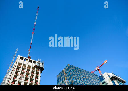 Niedrigen Winkel Blick auf zwei konkrete Gebäude im Bau mit Turm Kräne an ihrer Spitze neben einem Glasbau gegen blauen Himmel. Stockfoto