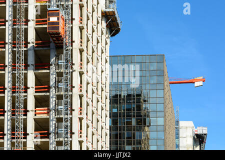 Vorderansicht des ein Glasgebäude zwischen zwei Betonbauten im Bau mit einem roten Turmdrehkran gegen blauen Himmel. Stockfoto