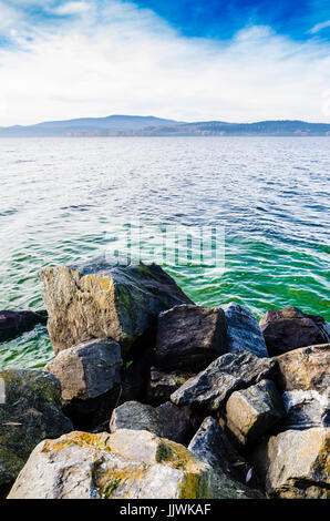 Einige Felsen vor schönen großen See mit Wellen. Auf der anderen Seite gibt es Wald, Berge und leichten Dunst. Herbst Wolken am blauen Himmel farbige. Stockfoto