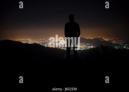 Silhouette eines jungen Mannes Stand oben auf dem Berg unter Nachthimmel mit Sternen und in der Nähe der Stadt. Stockfoto