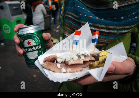 Amsterdam, Niederlande - 25. April 2017: Hände halten ein Bier Heineken und eine traditionelle holländische Delikatesse der Hering mit Gurken und Zwiebeln. Stockfoto