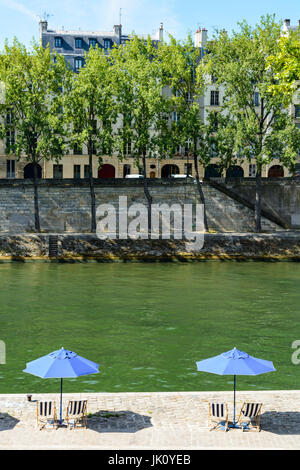 Zwei blaue Sonnenschirme mit blau-weiß gestreiften Liegestühlen in der Sonne am Ufer des Flusses Seine mit Pappeln und typisch Paris Gebäuden ich Stockfoto