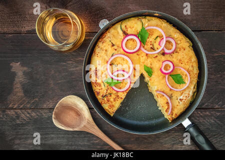 Eine obenliegende Foto eine spanische Tortilla mit Kartoffeln und Zucchini in eine Tortillera mit einem fehlenden Stück, mit einem Glas Weißwein und ein hölzernes Bursche Stockfoto